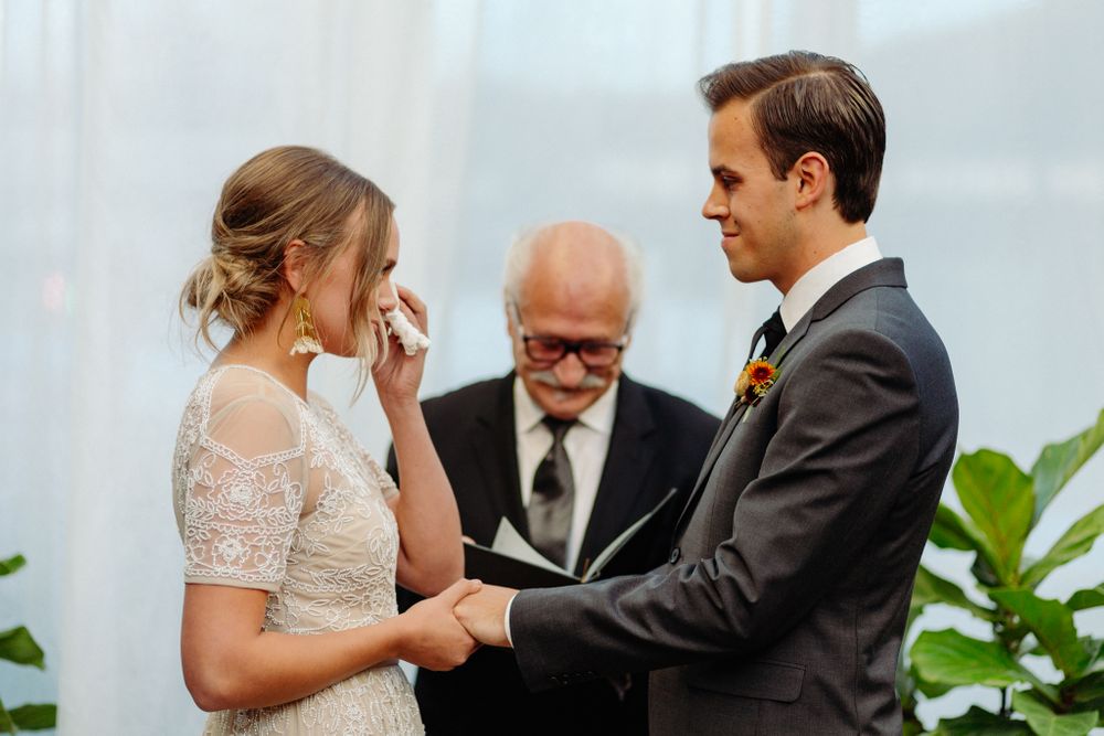 An emotional moment during a wedding ceremony in front of sheer white curtains and tropical plants.