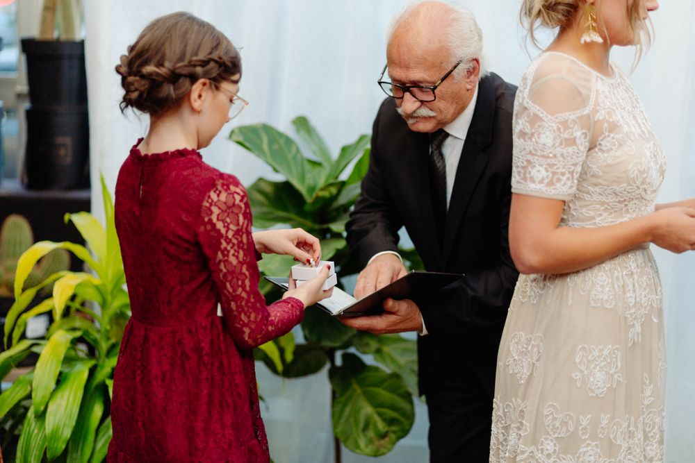 Wedding participants exchange rings during an indoor ceremony surrounded by tropical plants.