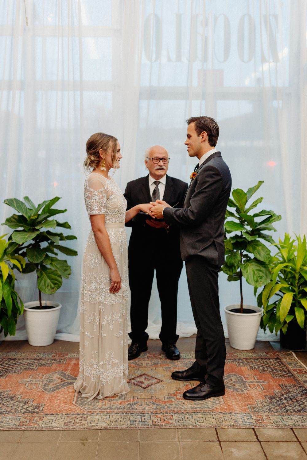 A wedding ceremony in progress on a brick floor with white curtains and potted plants as backdrop.