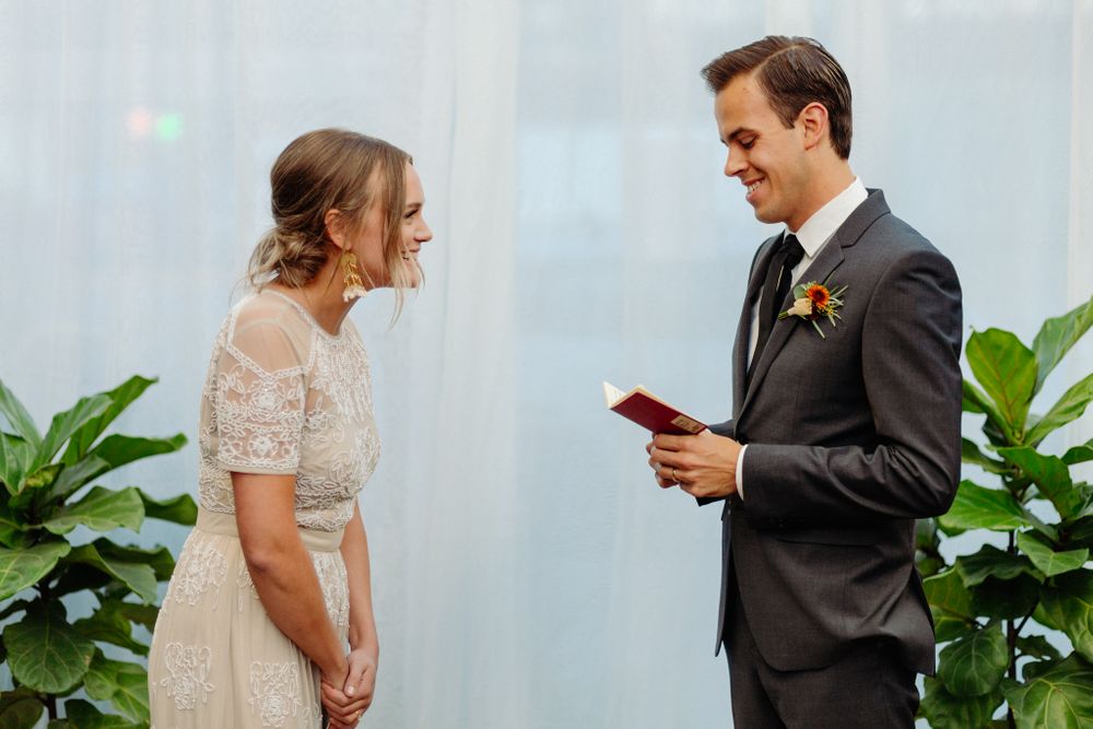 Couple exchanging vows during wedding ceremony with white backdrop and potted plants.