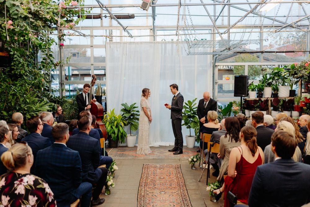 Wedding ceremony in greenhouse with guests seated on both sides of oriental rug aisle.