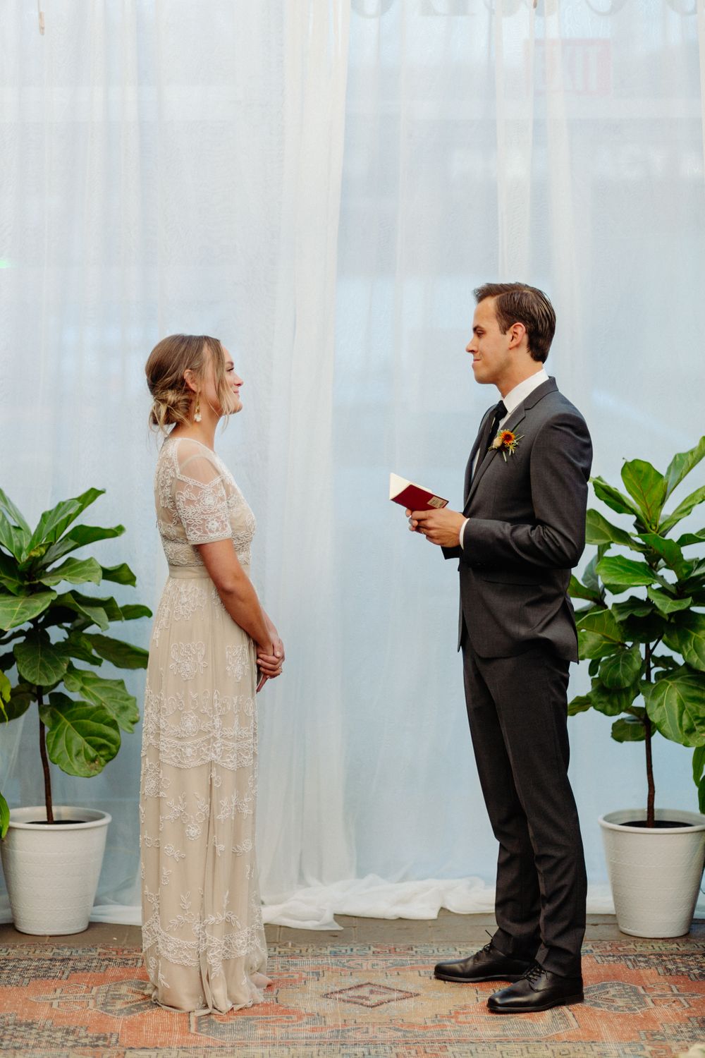 Wedding ceremony with couple standing between potted plants against white backdrop.