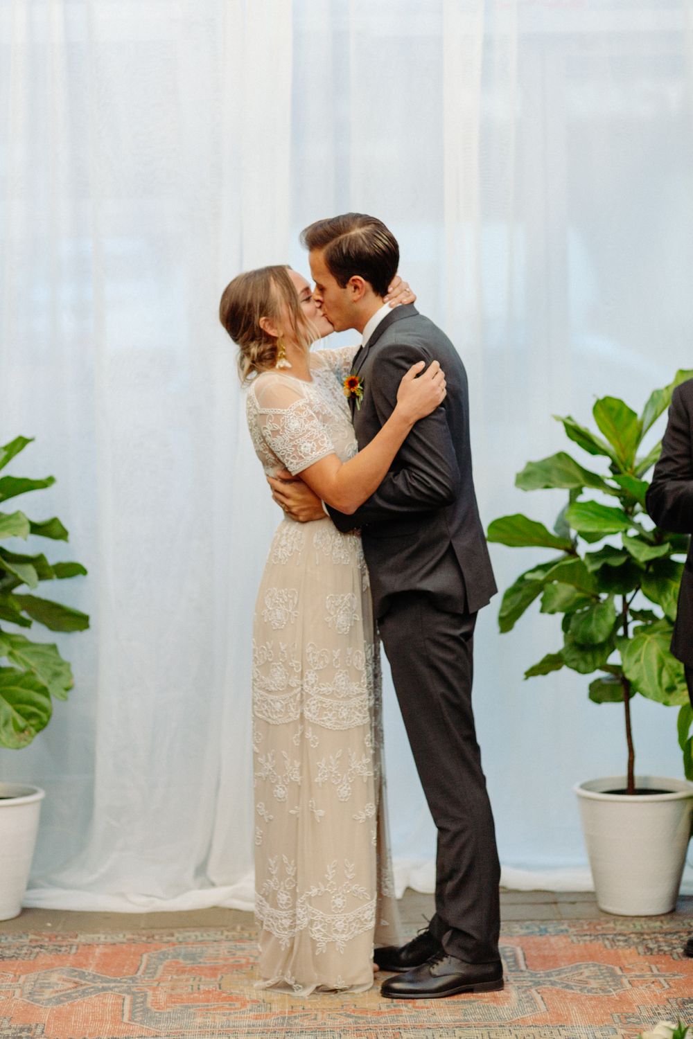 Couple shares romantic kiss in front of white curtain backdrop with potted plants at intimate wedding ceremony.