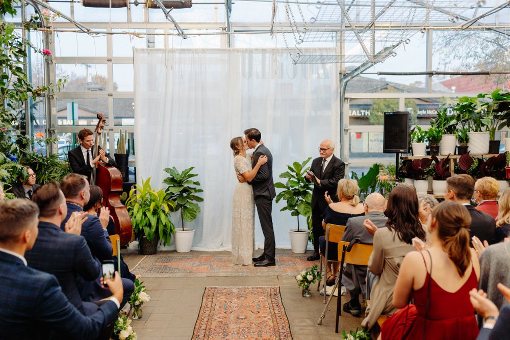 Wedding ceremony in greenhouse with white curtain backdrop and guests seated on vintage chairs.