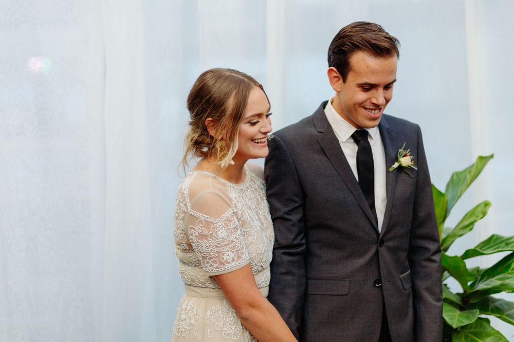 Newlyweds share joyful moment in front of white backdrop with greenery.