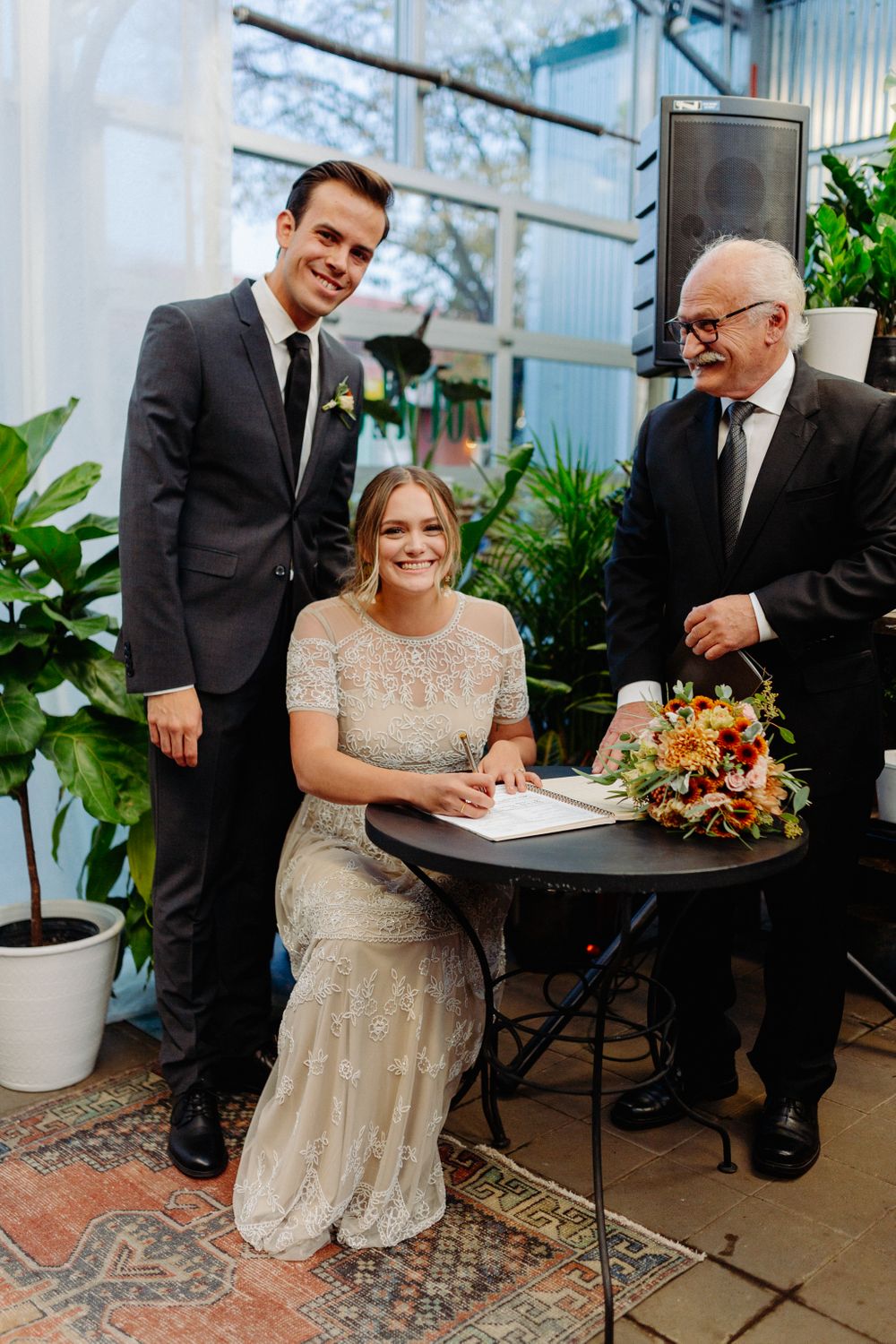 Wedding party gathers around small table for marriage license signing in greenhouse setting.