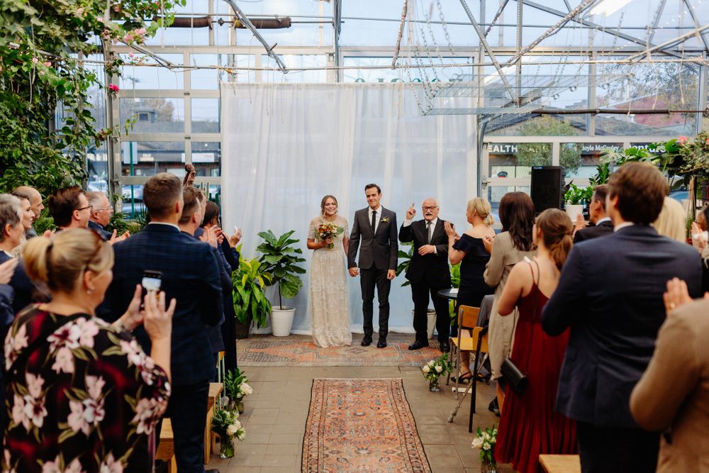 Wedding guests capture photos of ceremony recessional in greenhouse setting with vintage decor.
