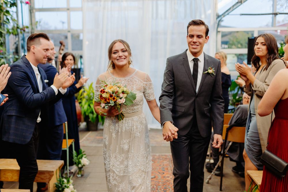 Newlyweds walk down aisle while wedding guests applaud in greenhouse venue.