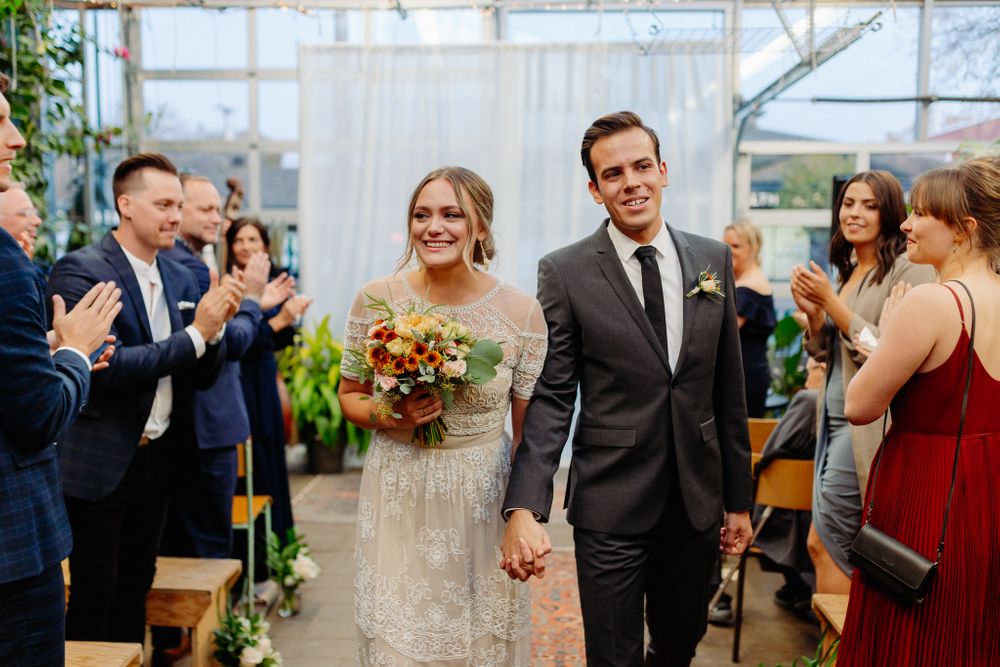 Newlyweds walk down the aisle in a greenhouse wedding venue while guests applaud their joyful celebration.