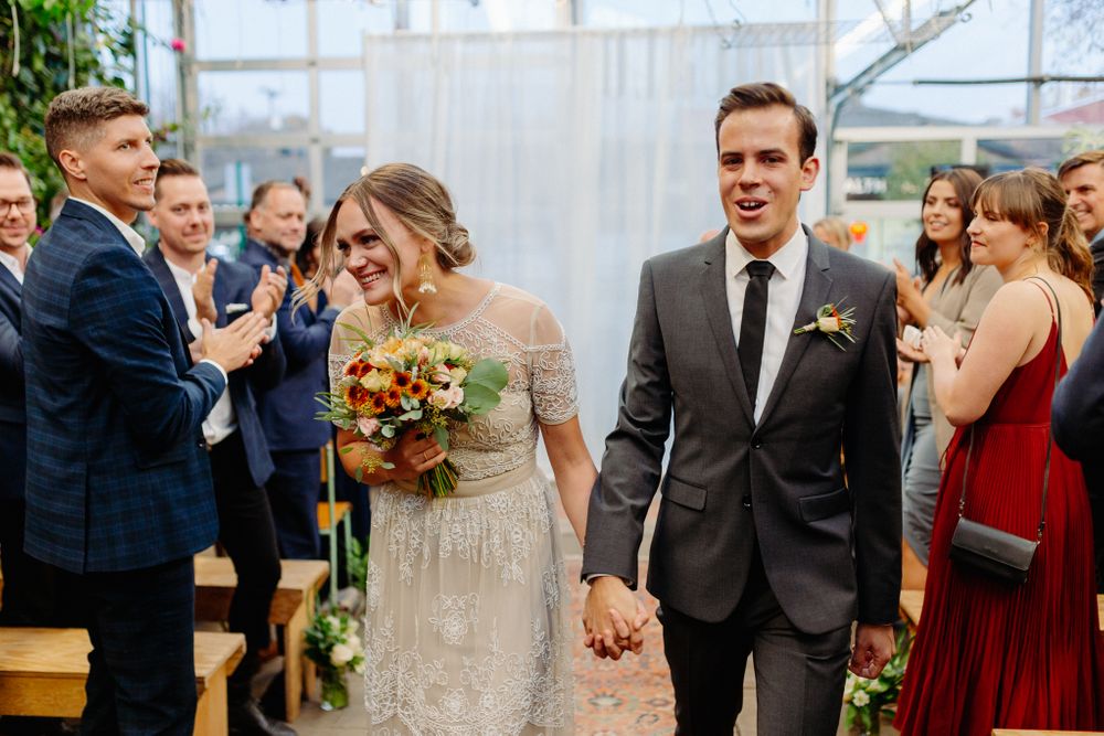 A wedding couple walks hand in hand through a glass conservatory as guests clap during the recessional.