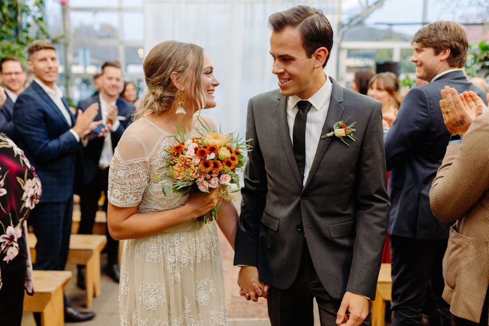 Wedding couple exchanges loving glances while holding autumn-colored bouquet in greenhouse setting.