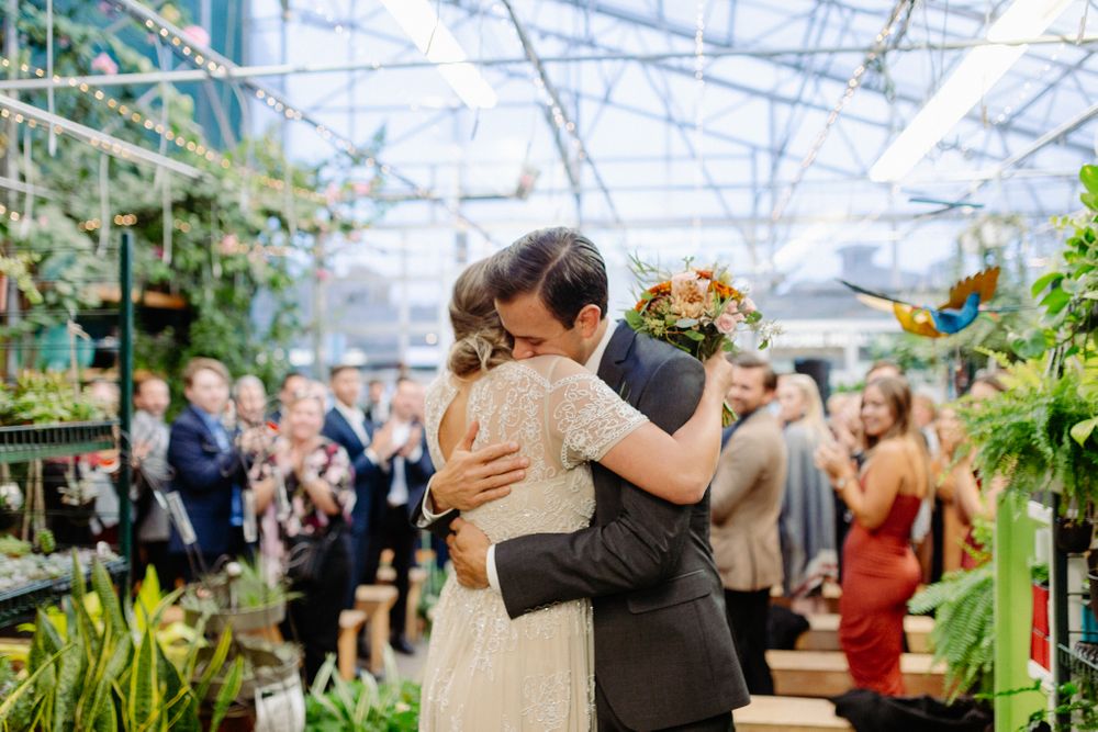 Couple embraces during emotional wedding ceremony moment in a sunlit greenhouse surrounded by plants.
