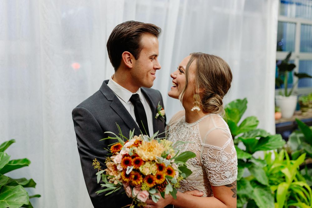 Joyful couple laughs together while holding vibrant fall wedding bouquet in greenhouse setting.
