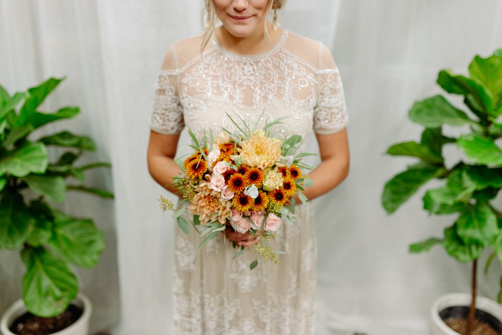 Elegant lace wedding dress with delicate sleeves and a vibrant fall-colored bouquet against a white backdrop.