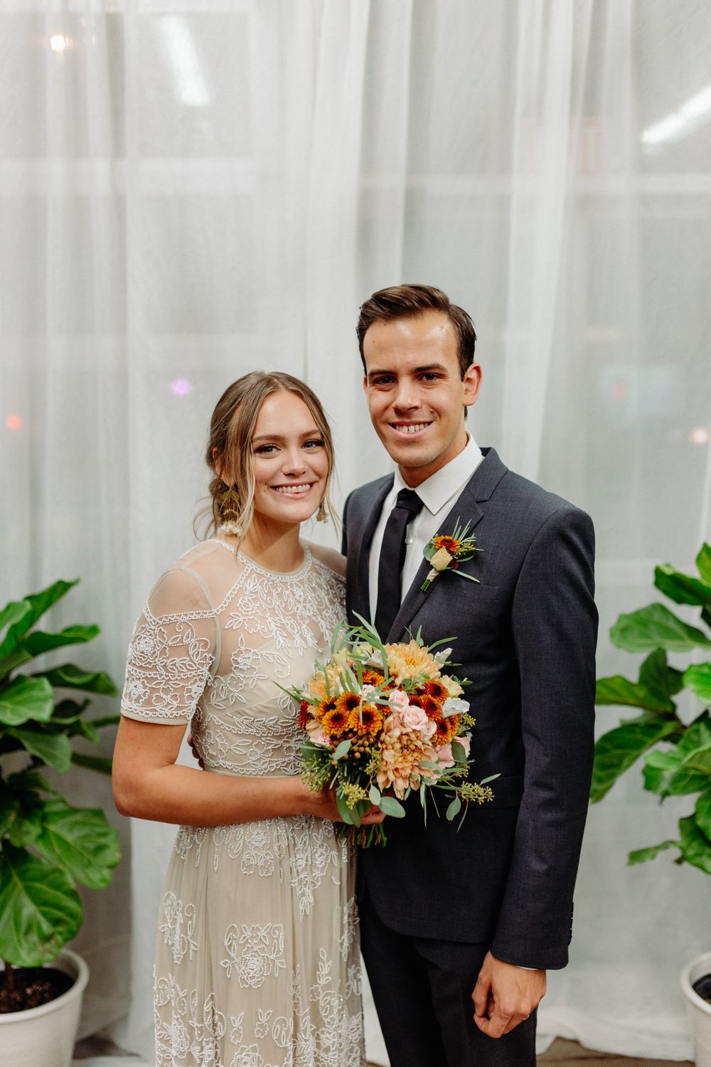 Wedding couple in a lace dress and dark suit pose with a colorful fall bouquet against white curtains.