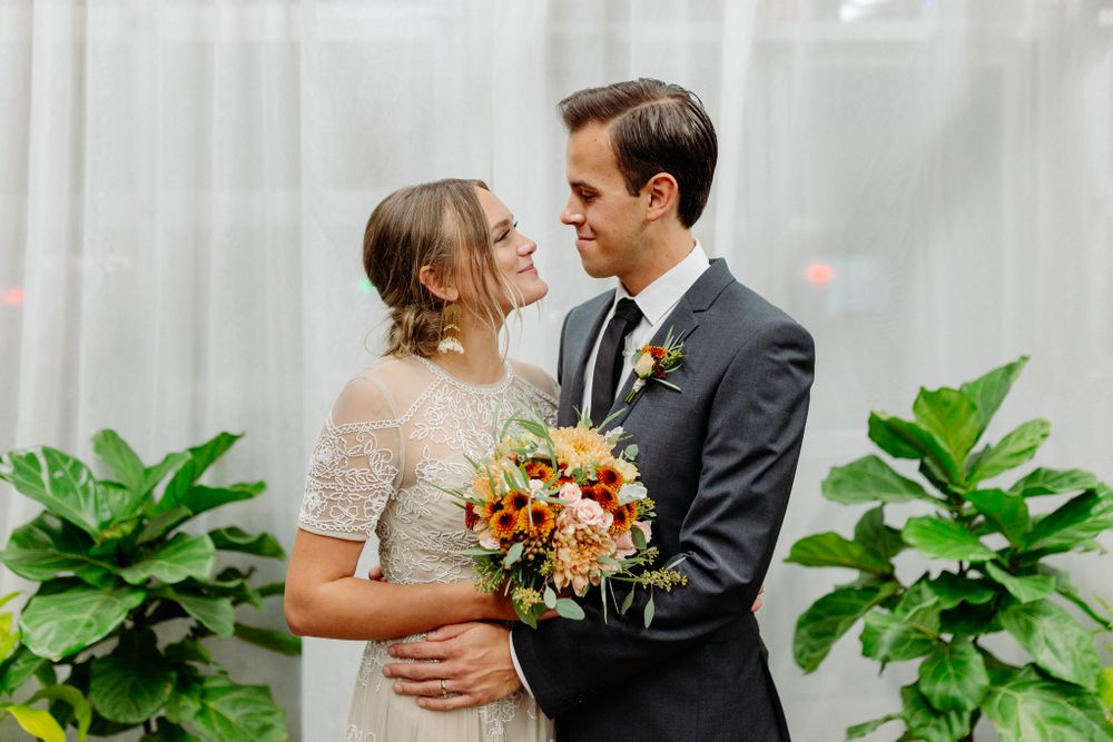 Intimate wedding portrait against white curtains with lush green plants and warm autumn bouquet.