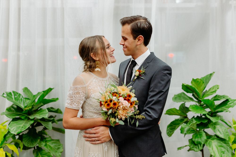 Romantic wedding moment captured against white curtains and green plants with fall-colored florals.