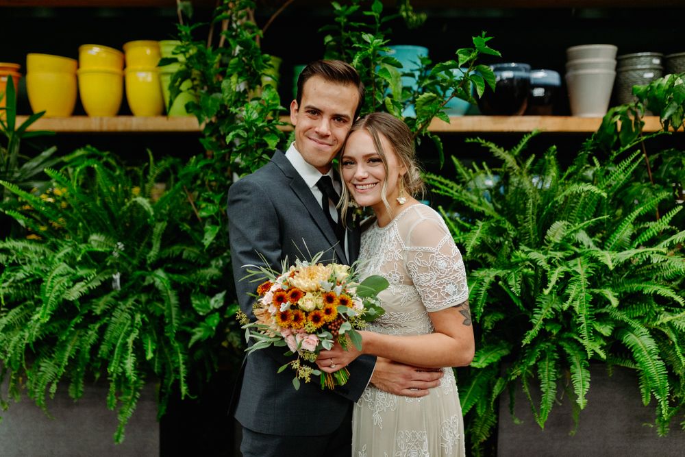 Wedding couple poses in front of lush green plants and yellow pots.
