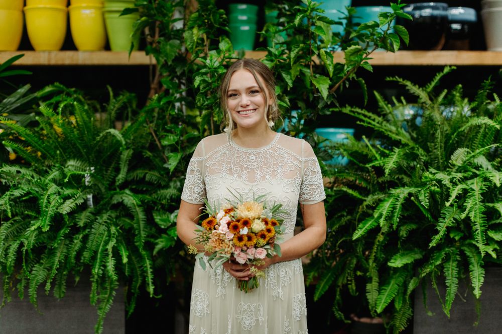 Bride in beaded ivory dress holds autumn-colored bouquet against fern wall.