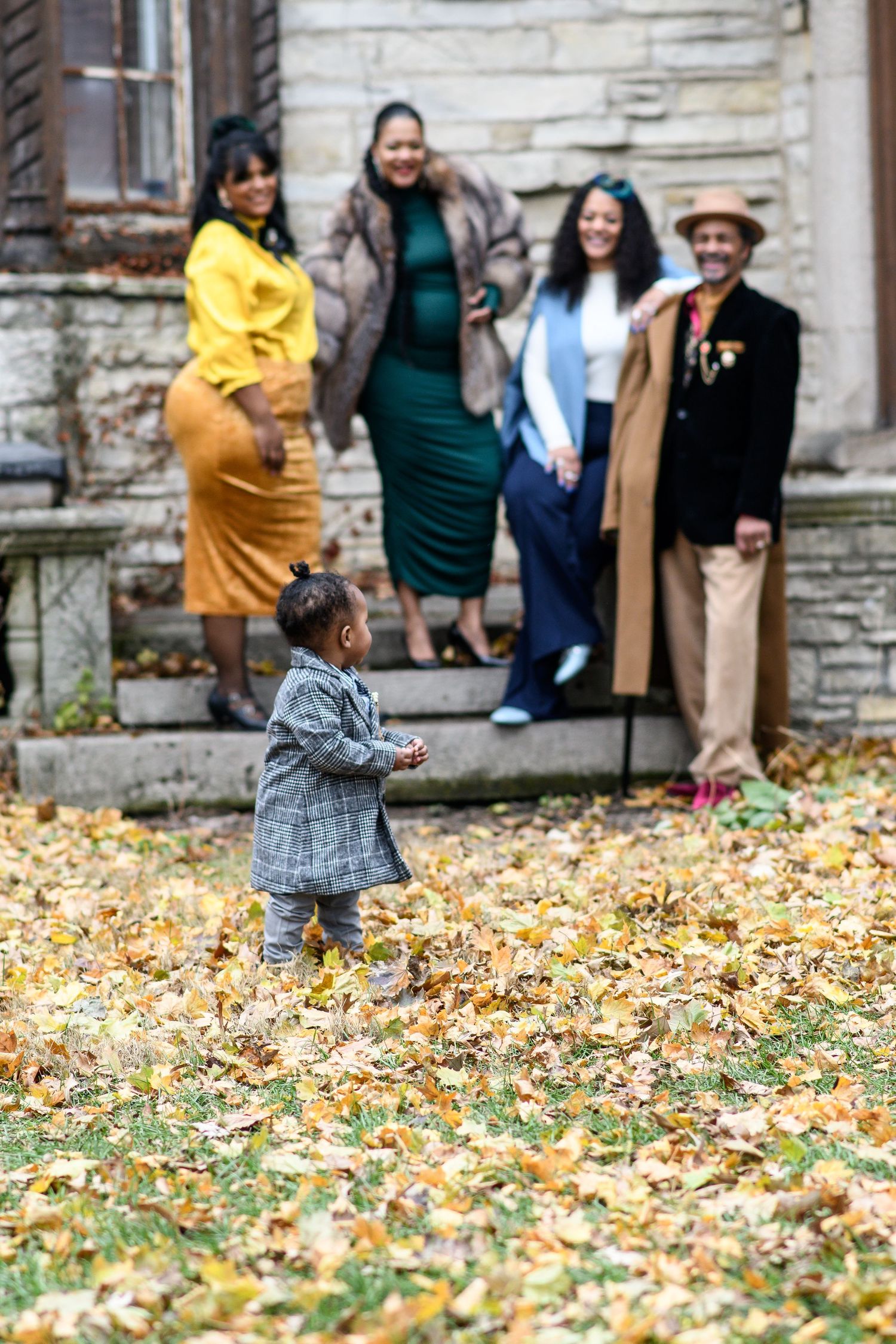 Child running through fallen autumn leaves while group watches from stone building background.