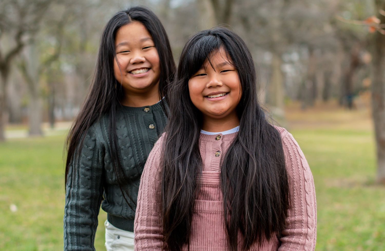 Smiling siblings sit together on ground wearing casual outfits in natural outdoor setting.