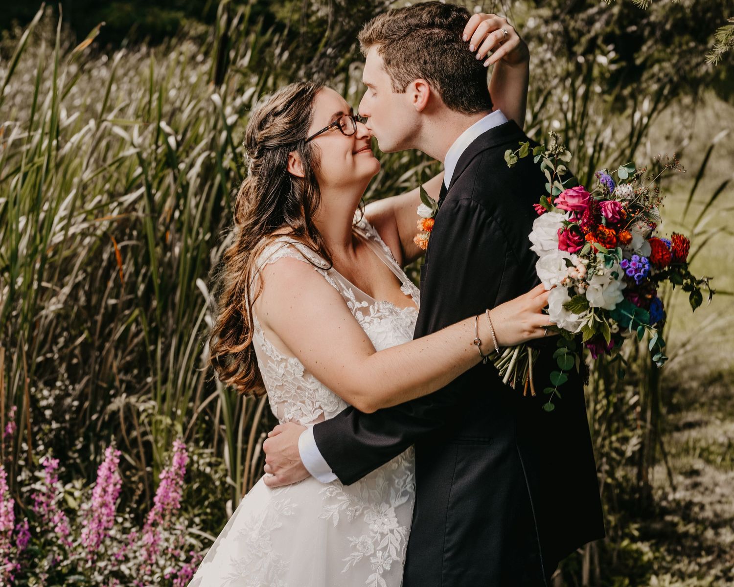 Newlyweds kiss in garden setting with colorful bouquet of flowers.