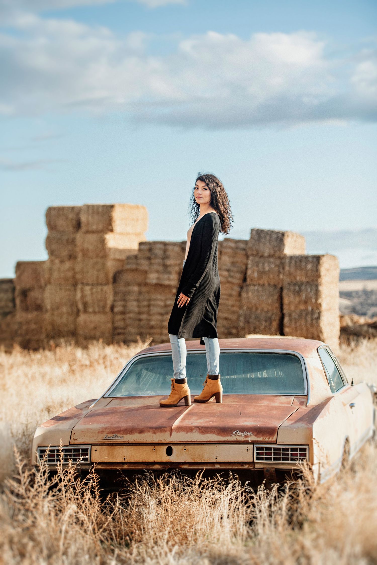 Senior girl stands on an old car near a haystack