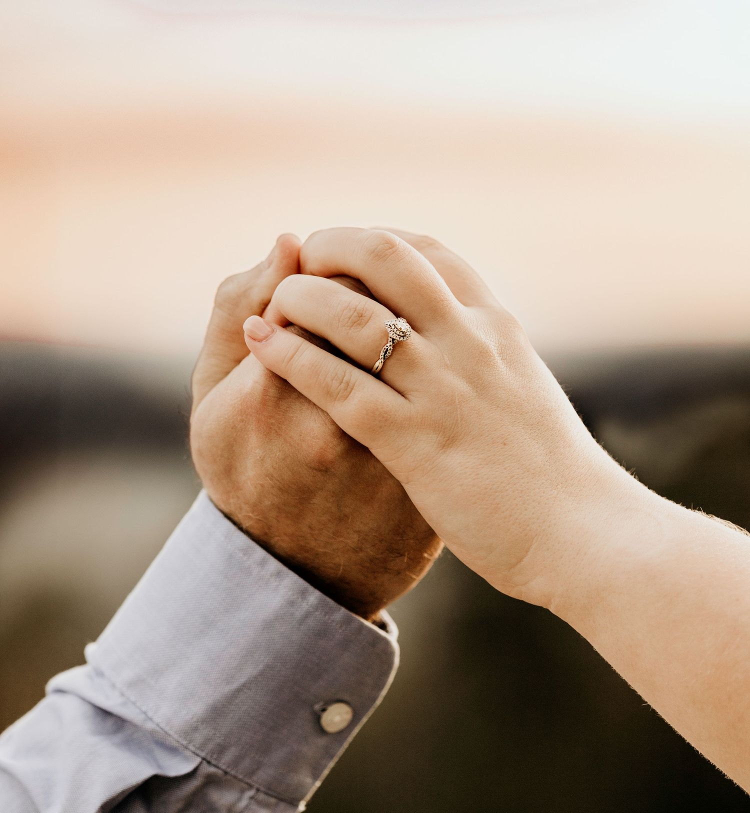 Two hands clasped together with engagement ring visible, against soft sunset background.