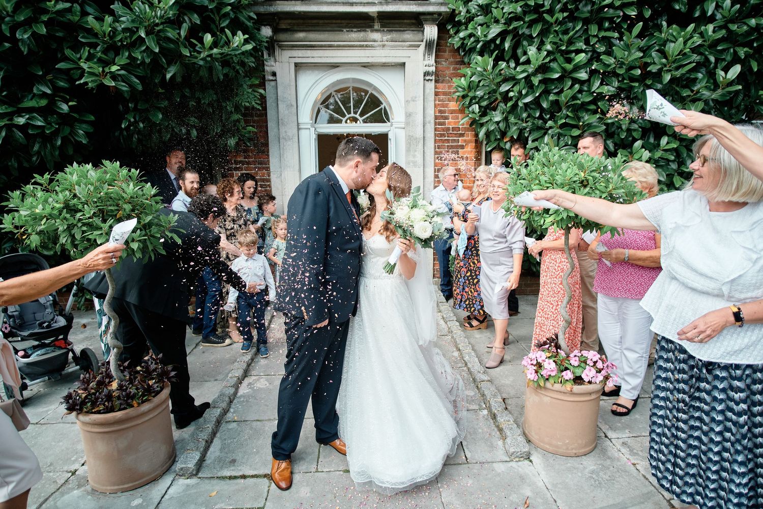 Newlyweds share kiss as guests throw confetti outside historic venue entrance.