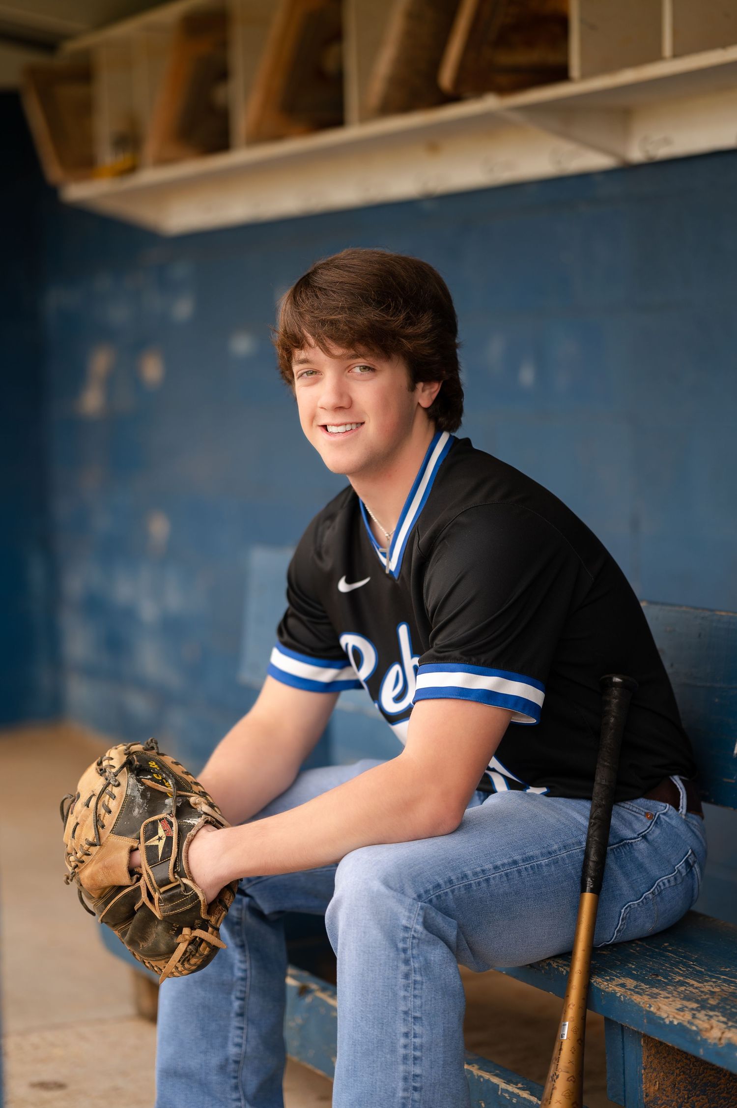 Baseball player in black uniform sitting in dugout with glove and bat nearby.