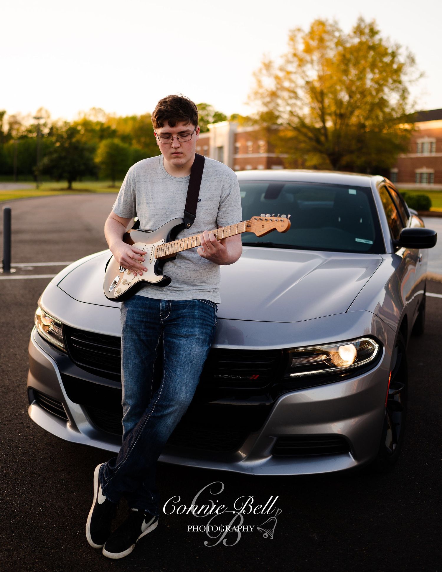 Person playing electric guitar while sitting on hood of silver Dodge Charger in parking lot.