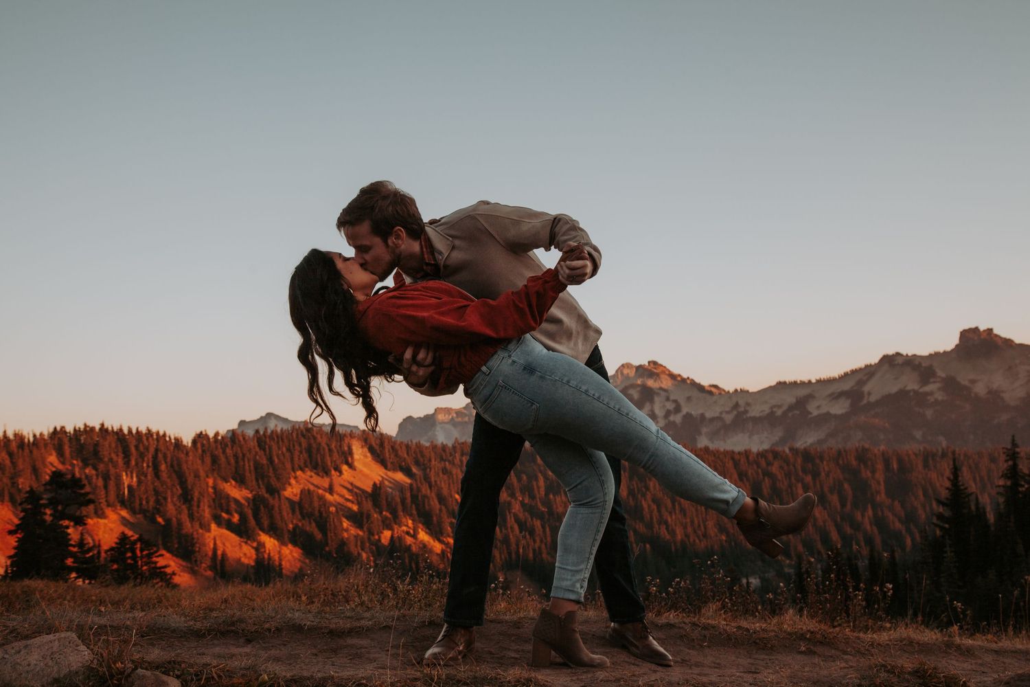 Couple shares a romantic dip kiss on a mountain overlook with autumn-colored hills and evergreen trees in the background.