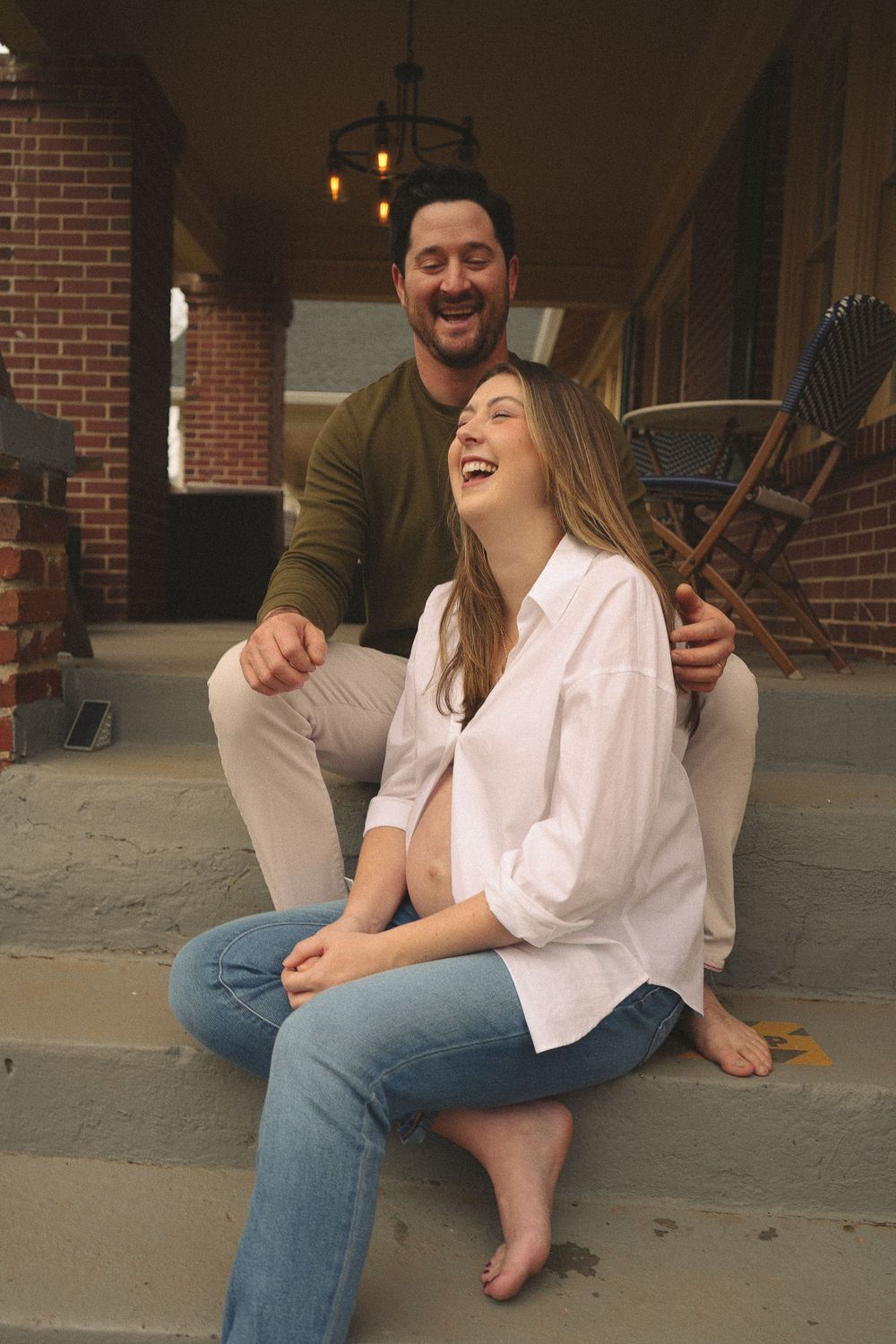 Happy pregnant couple laughing together on front porch steps of brick home.