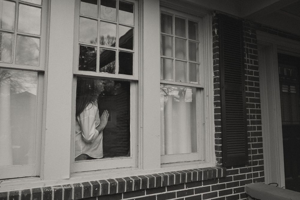 Black and white photo of person visible through open window of brick house exterior.
