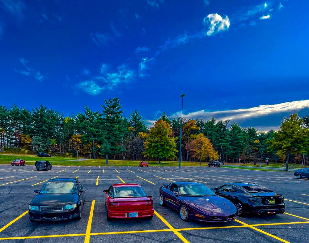 Multiple luxury cars parked in a lot under bright blue skies with trees visible in the background.
