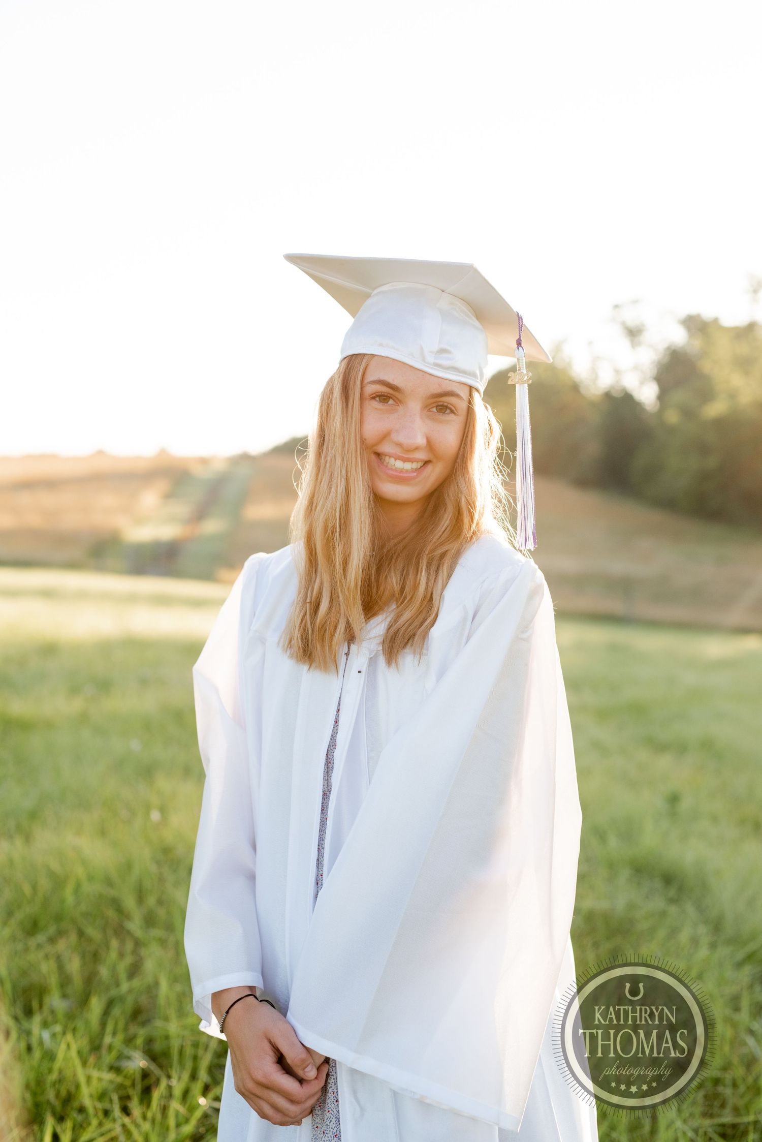 Graduate in white cap and gown poses in golden sunlit field during sunset graduation photoshoot.