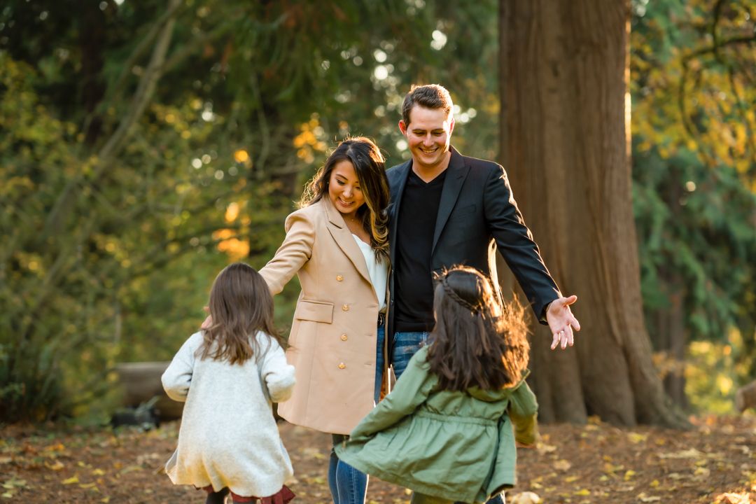Family plays and walks together under large trees with golden autumn leaves.