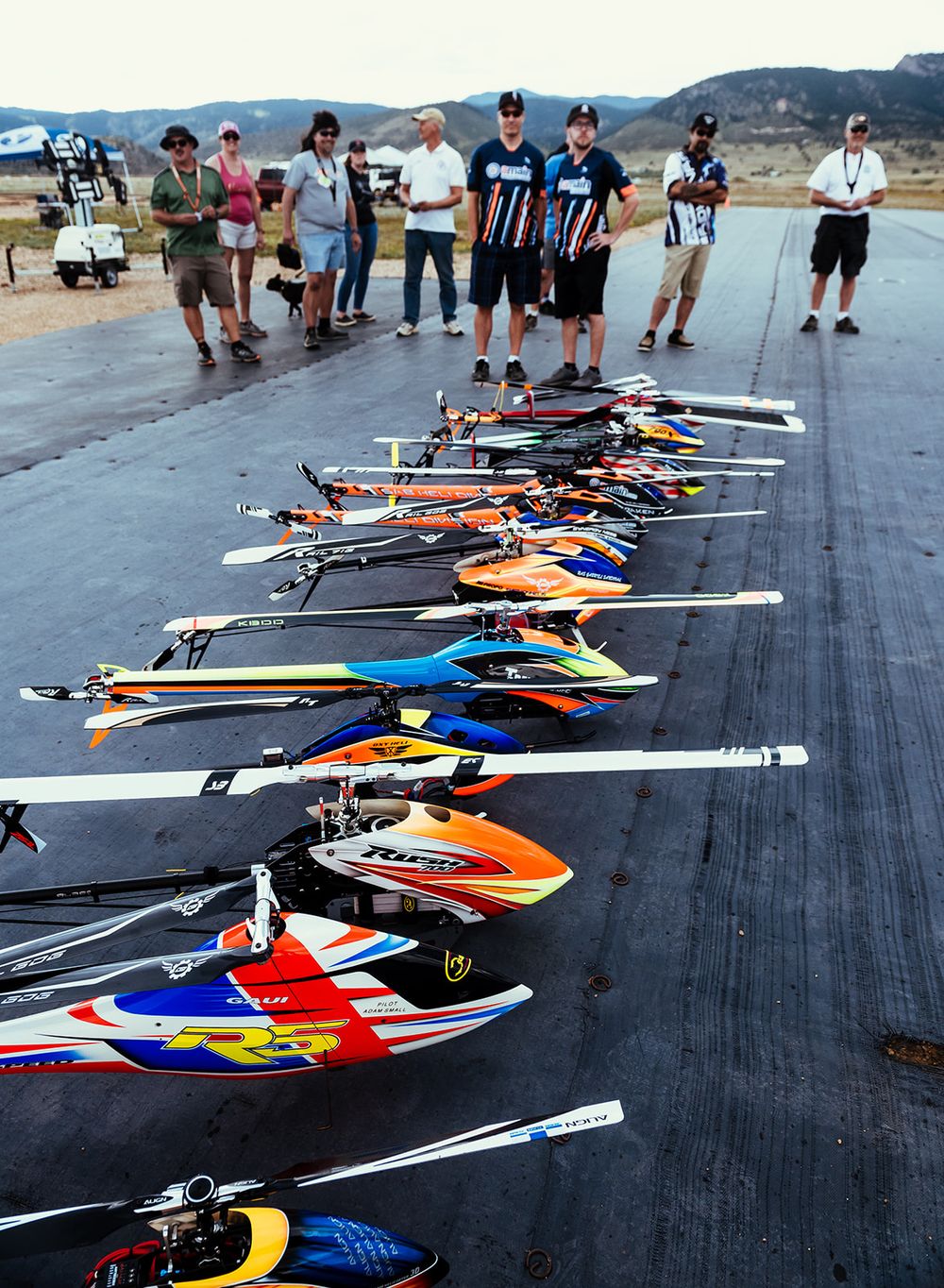 Row of colorful RC helicopters lined up on pavement with spectators watching in background.