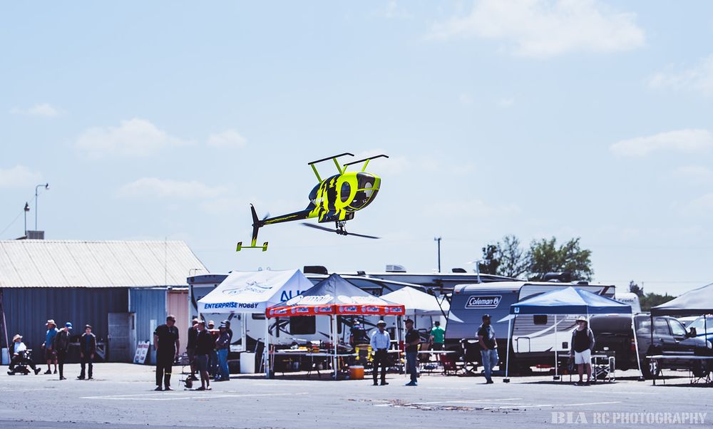 Yellow RC helicopter performing aerial maneuver above event tents and spectators.