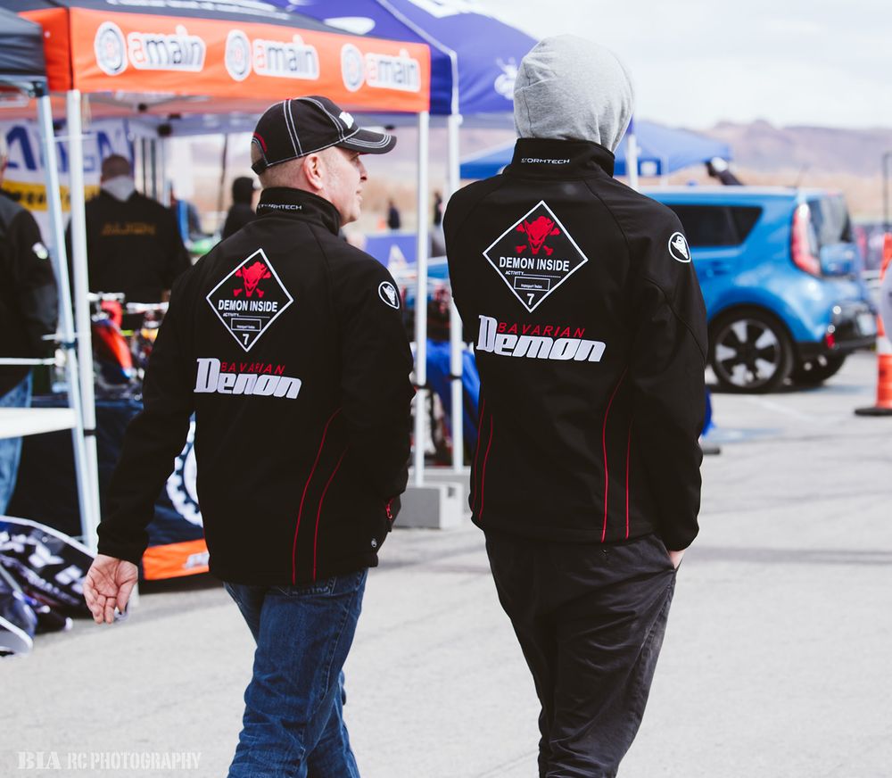 Two people wearing black Lehman RC team jackets stand at an outdoor event with orange tents in the background.