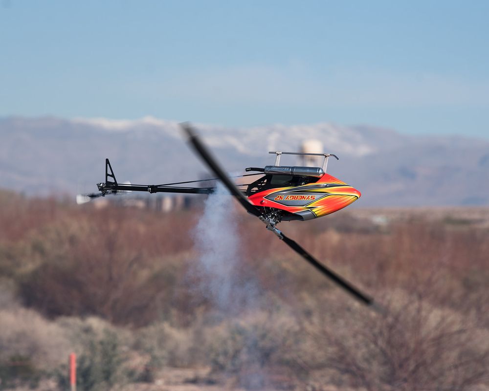Remote control helicopter performing an aerobatic maneuver against a desert mountain backdrop.