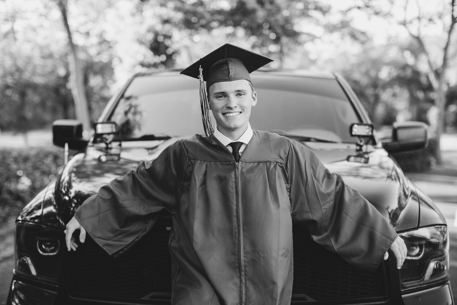 Graduate in cap and gown poses with arms outstretched in front of car.