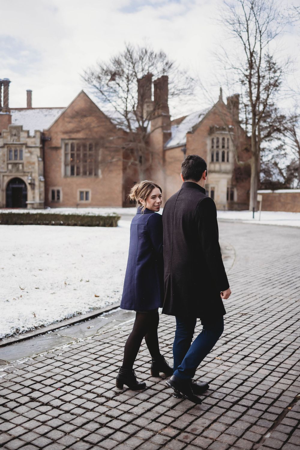 A couple walks together on snow-covered brick ground near historic buildings.