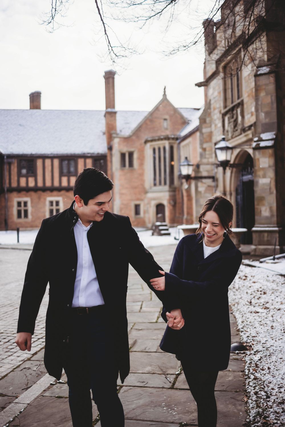 Playful couple runs and dances on snowy grounds of a brick university campus.