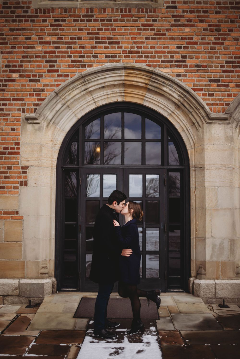 A romantic winter embrace under an arched brick doorway covered in light snow.