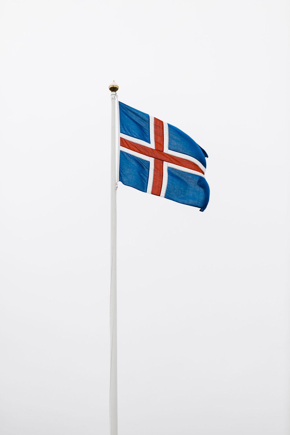 The Icelandic flag waves against an overcast sky on a white pole.