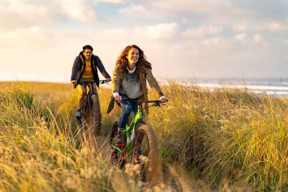 Cyclists riding through tall grass on coastal trail during golden hour sunset.