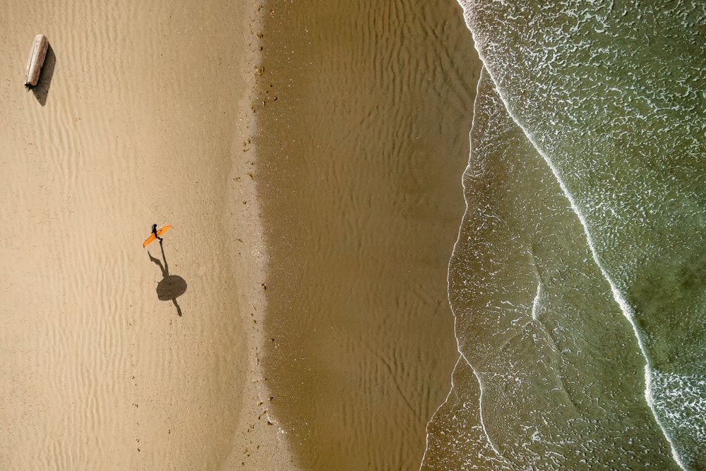 Aerial view of a lone figure walking along a sandy beach next to ocean waves.