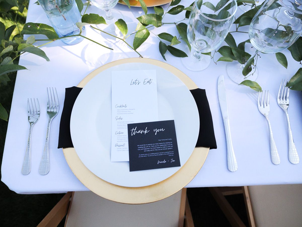 Elegant table setting with white plate, gold charger, black thank you card and silverware under green foliage.