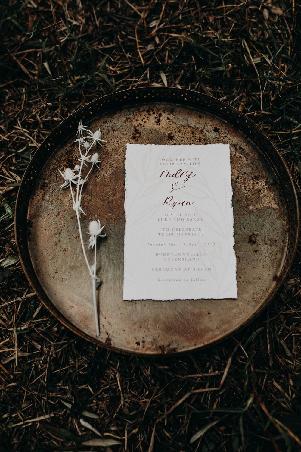 Wedding invitation with delicate dried flowers on a rustic metal plate against dark soil.